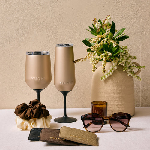 Two beige wine glasses with a vase of flowers and sunglasses on a light surface.