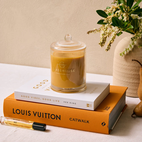 Candle in a glass jar on top of a Louis Vuitton book with a vase of flowers in the background.