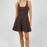 A woman modeling a sleeveless, scoop-neck mini dress in a dark brown color, paired with black flip-flop sandals, against a light gray studio background.