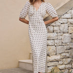 Woman in a white dress with black polka dots standing on a stone path next to a stone wall.