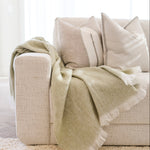 A light sage green throw blanket with white fringe is draped over a beige fabric sofa with neutral pillows in a bright, modern living room.