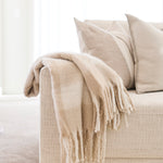 A cozy beige throw blanket with fringe is draped over the arm of a light beige upholstered sofa, alongside matching pillows in a bright, minimalist room.