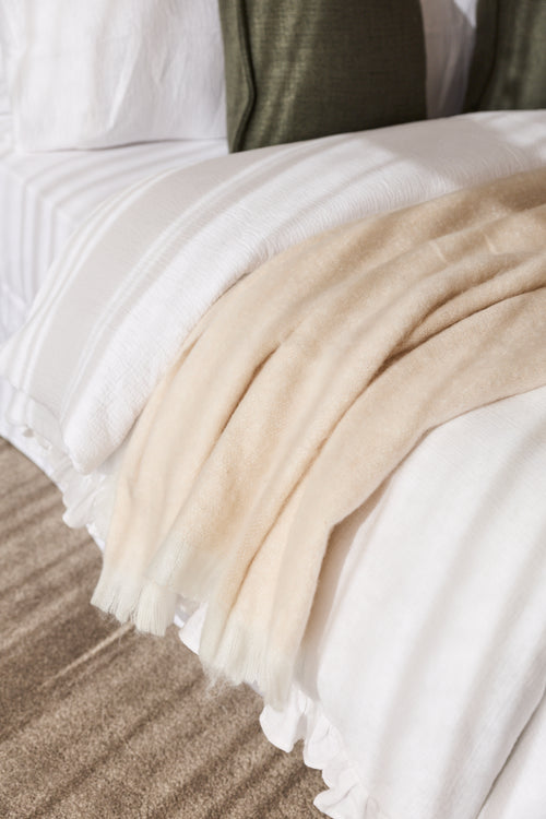 A close-up of a soft cream throw blanket draped over white bedding on a bed, with a textured taupe rug visible at the bottom.