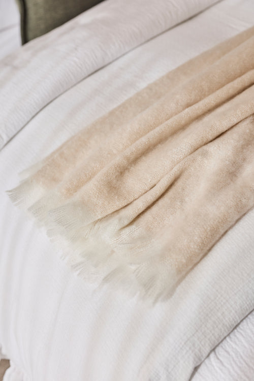 A close-up of a soft, light beige throw blanket with fringed edges draped over white textured bedding.