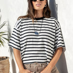 A woman wearing a black and white horizontally striped short-sleeve t-shirt, beige cargo pants, and black sunglasses, leaning against a white stucco wall outdoors.