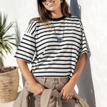 A woman wearing a black and white horizontally striped t-shirt, beige wide-leg pants, and black sunglasses, leaning against a white stucco wall outdoors.