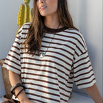 A woman wearing a brown and cream striped short-sleeve t-shirt and a dark brown skirt, sitting on a white ledge in a sunlit minimalist setting with a cactus in the background.