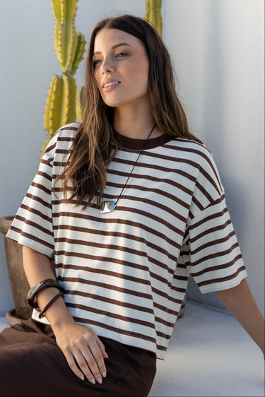 A woman wearing a brown and cream striped short-sleeve t-shirt and a dark brown skirt, sitting on a white ledge in a sunlit minimalist setting with a cactus in the background.