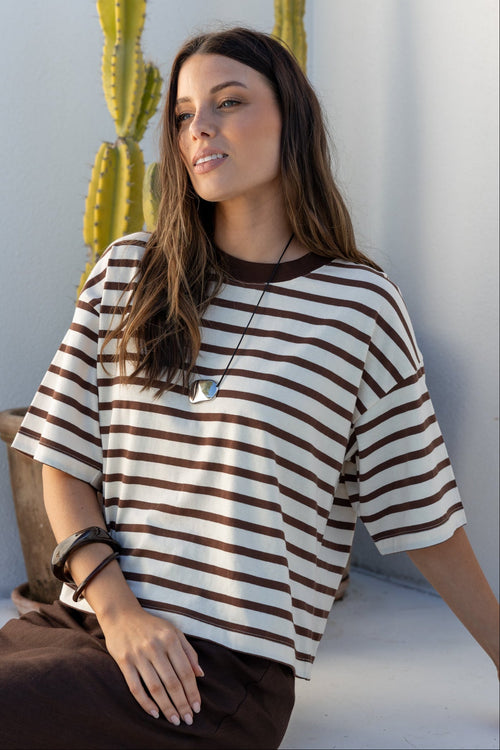 A woman wearing a brown and cream striped short-sleeve t-shirt and a dark brown skirt, sitting on a white ledge in a sunlit minimalist setting with a cactus in the background.