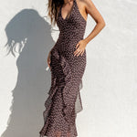 A woman modeling a dark brown sleeveless maxi dress with a white floral pattern, standing outdoors against a white stucco wall with a pool in the foreground.