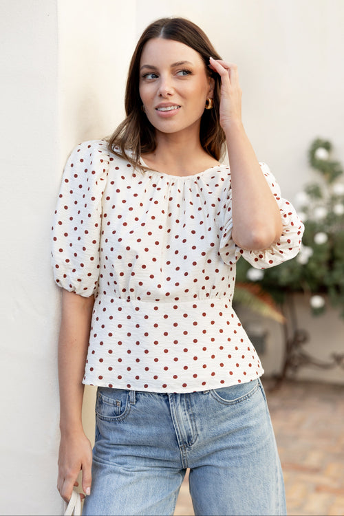 A woman wearing a white short-sleeve blouse with brown polka dots and light blue jeans, standing against a light-colored wall.