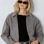 Woman wearing a gray jacket, black top, and blue jeans against a white wall.