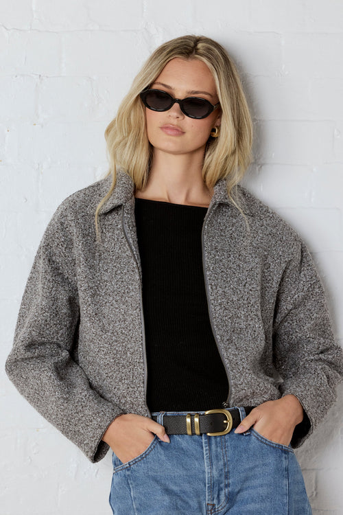 Woman wearing a gray jacket, black top, and blue jeans against a white wall.