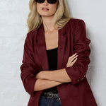 Person wearing a burgundy blazer and sunglasses against a white brick wall