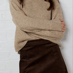 Person wearing a beige sweater and brown skirt against a white brick wall.