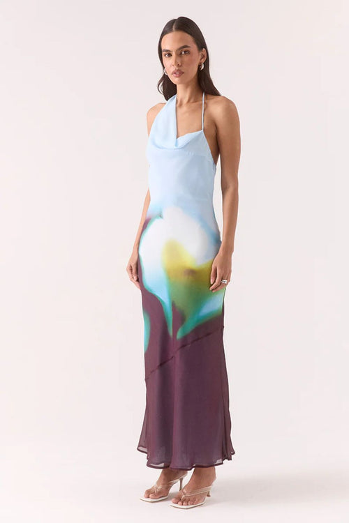 Model wears halter-neck maxi dress with blurred azure, green, and plum abstract floral print, asymmetric hem, and a softly draped neckline.