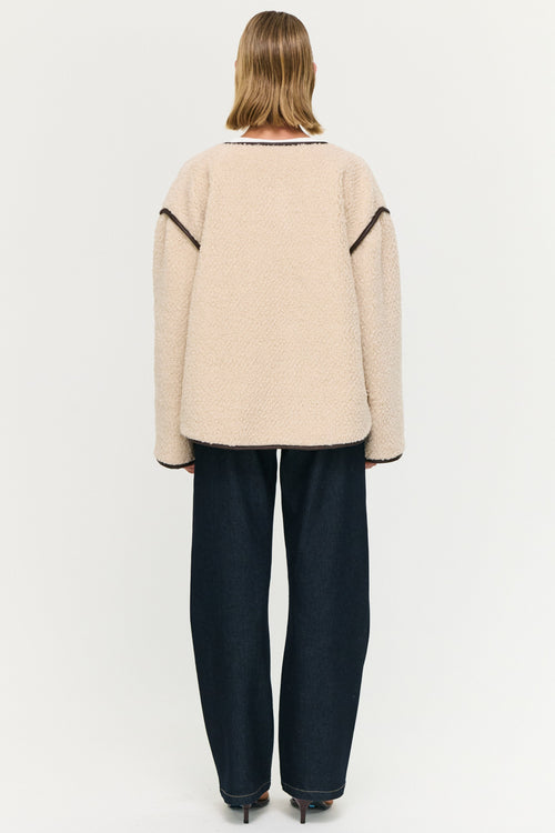 Person wearing a beige sweater with black trim and dark pants on a white background