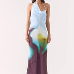 Model wears halter-neck maxi dress with blurred azure, green, and plum abstract floral print, asymmetric hem, and a softly draped neckline.