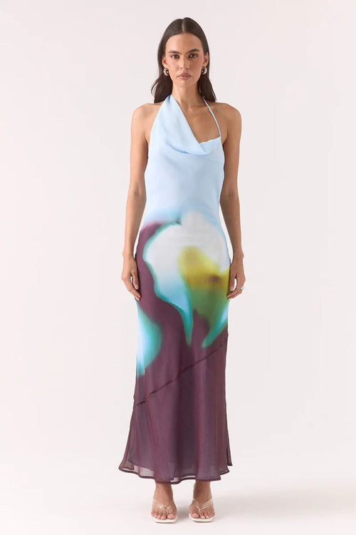 Model wears halter-neck maxi dress with blurred azure, green, and plum abstract floral print, asymmetric hem, and a softly draped neckline.