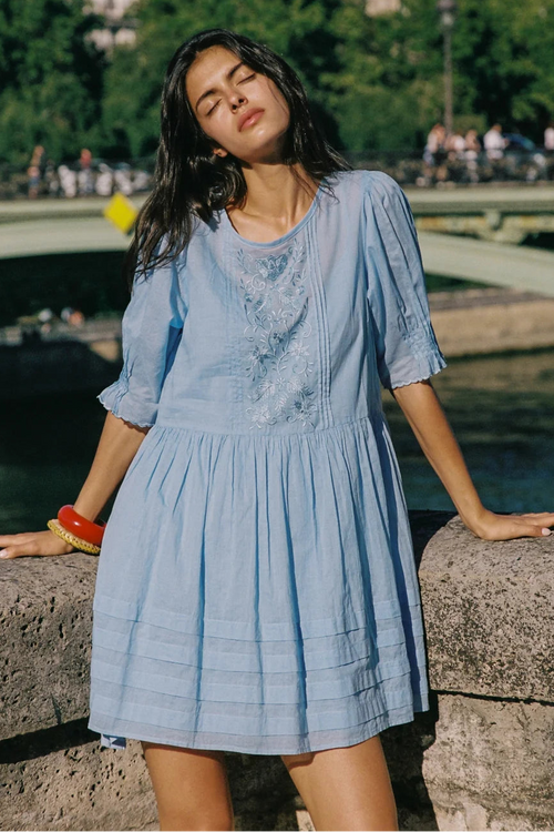 A woman wearing a light blue cotton dress with embroidered details on the chest, leaning on a stone bridge with a river and green trees in the background.