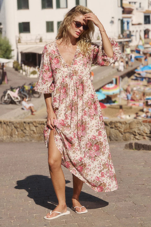 A woman wearing a long-sleeve, V-neck maxi dress with a pink and green floral print on a cream background, standing outdoors on a sunny day with a beach town background.