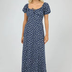 A woman modeling a navy blue maxi dress with a small white floral print, puff sleeves, and a scoop neckline with a tie detail at the front, set against a plain light gray studio background.