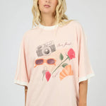A woman modeling an oversized light pink t-shirt with a graphic print of a camera, sunglasses, a strawberry, and flowers, paired with blue jeans against a light gray background.