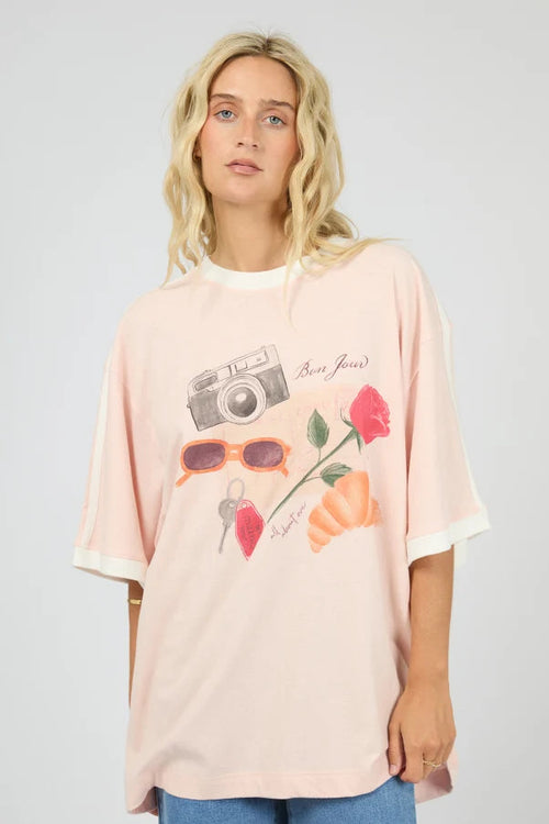A woman modeling an oversized light pink t-shirt with a graphic print of a camera, sunglasses, a strawberry, and flowers, paired with blue jeans against a light gray background.