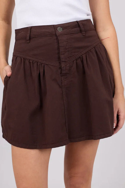 A dark brown high-waisted mini skirt with a pleated front and side pockets, shown on a model wearing a white top against a light gray background.