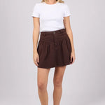 A woman modeling a white short-sleeve t-shirt and a dark brown pleated mini skirt, paired with black flip-flop sandals, against a light gray studio background.