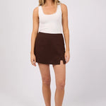 A woman modeling a white sleeveless top with scalloped edges and a dark brown mini skirt, paired with black flip-flop sandals, against a light gray studio background.