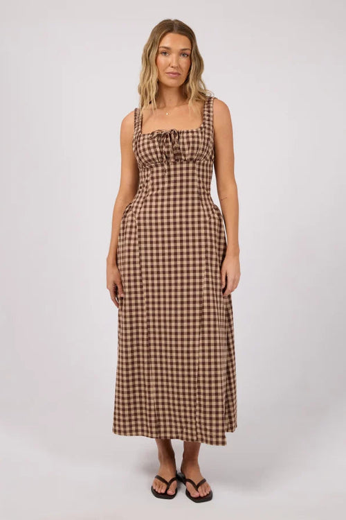 A woman modeling a brown and beige gingham maxi dress with a tie-front bodice, photographed against a plain light gray studio background.