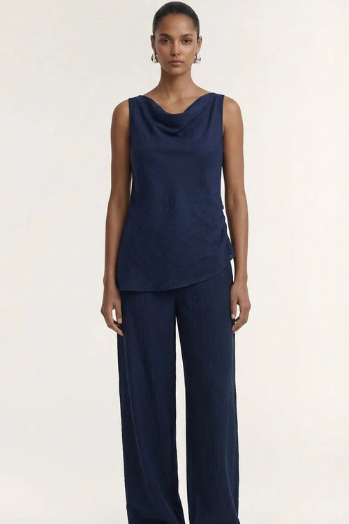 A model stands against a plain light beige studio background wearing a dark navy sleeveless draped top and matching wide-leg pants, with brown sandals.
