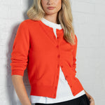 A blonde woman wearing a bright orange cardigan over a white top and black trousers, leaning against a white brick wall.
