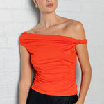 A woman modeling a bright orange off-the-shoulder ruched top paired with black pants, standing against a white painted brick wall.
