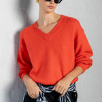 A woman wearing a bright orange-red V-neck knit sweater, black sunglasses, and a black skirt with a navy and white striped shirt tied at the waist, posing against a white brick wall.