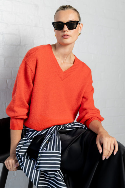 A woman wearing a bright orange-red V-neck knit sweater, black sunglasses, and a black-and-white striped shirt tied around her waist, seated against a white brick wall.