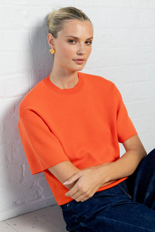 A woman wearing a bright orange short-sleeve crewneck t-shirt and dark blue jeans, sitting against a white brick wall.