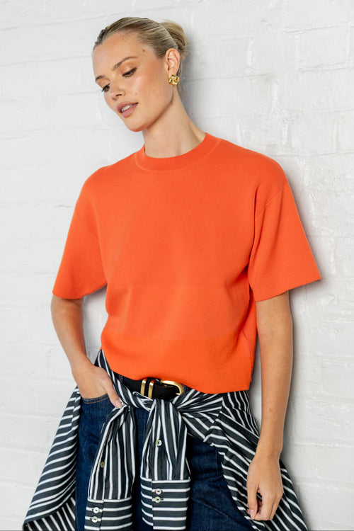 A woman wearing a bright orange short-sleeve crewneck knit top, styled with a navy and white striped shirt tied at the waist and dark blue jeans, posing against a white brick wall.