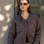 Woman wearing a brown quilted jacket and sunglasses standing against a neutral wall.