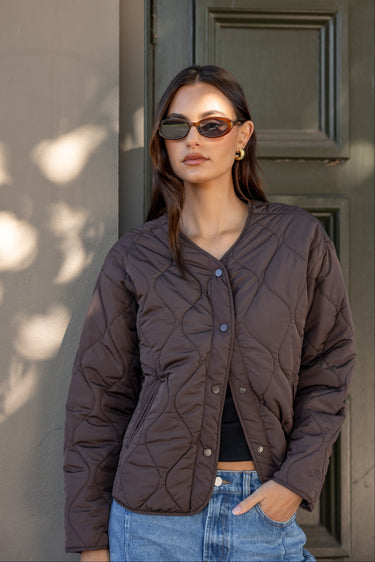 Woman wearing a brown quilted jacket and sunglasses standing against a neutral wall.