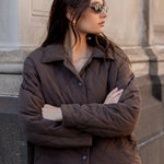 Woman wearing sunglasses and a brown jacket standing against a stone wall.