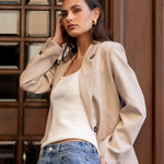 Woman wearing a beige blazer and blue jeans standing against a wooden paneled wall.