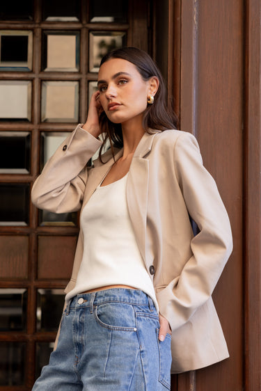 Woman wearing a beige blazer and blue jeans standing against a wooden paneled wall.