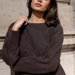 Woman wearing a dark brown sweater and blue jeans standing against a stone wall.