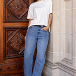 Woman in a white top and blue jeans standing against a stone wall with a wooden door.