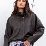 Woman wearing a dark jacket, striped shirt, and blue jeans against a white wall.