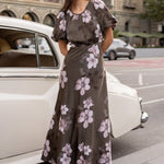 Woman in a floral dress standing next to a vintage car on a city street.