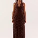 A woman modeling a dark brown halter-neck maxi dress with a subtle polka dot pattern, standing against a plain off-white studio background.