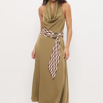 A woman modeling a sleeveless olive green halter maxi dress with a cowl neck, a matching tie belt, and a brown and white checkered patterned scarf tied at the waist, set against a plain white studio background.